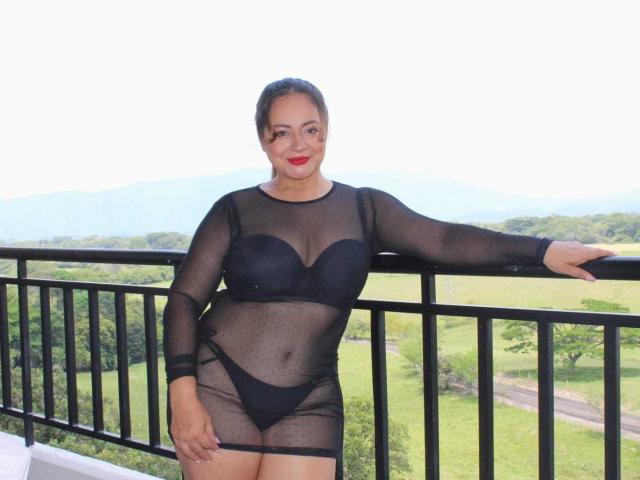 LolitaBusty with curvy body, medium skin, black bra, sheer black dress, and black panties, standing on balcony, left arm on rail, red lipstick, smiling.