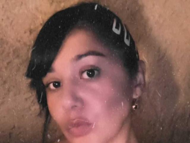 KikaSexy has dark hair with a white hairclip, brown eyes, and is pouting her lips. She's wearing small gold earrings and has a slightly pinkish skin tone.