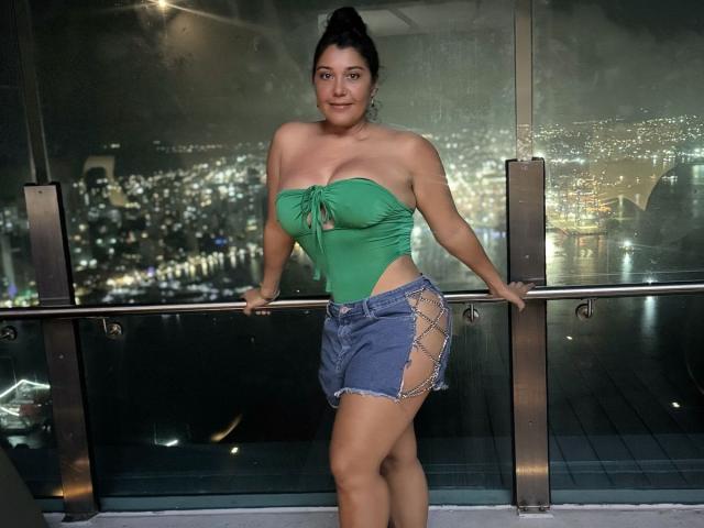 KikaSexy with big boobs in a green strapless top and blue denim shorts with lace-up sides, standing against a glass balcony at night, smiling.