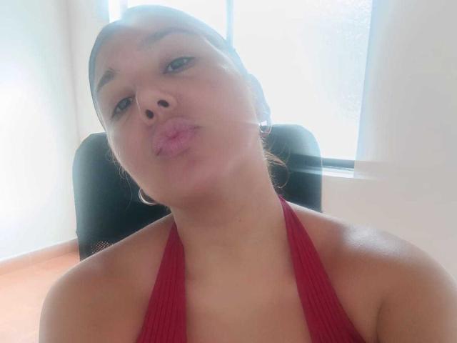 KikaSexy with pouty lips, brown hair in a bun, red halter top, and hoop earrings, her tits barely visible, standing in front of a black chair.