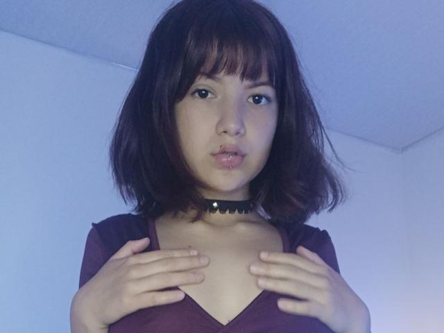 Caramelpearl has brown hair, a black choker, and a purple top, pouting with hands on her tits, in a blue-lit room. Her tits are small, hands cover them.