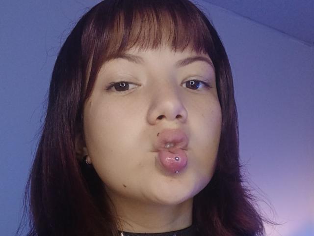 Caramelpearl has brown hair with bangs, brown eyes, and a lip piercing with a pink, heart-shaped, metal stud. She's pouting her full, slightly chubby, pink lips.