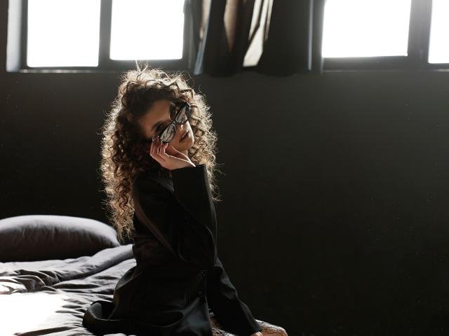 MiaAxel with curly brown hair, wearing black glasses and a black shirt, sitting on a bed, holding glasses to her face, sunlight streaming in from windows.