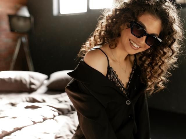 MiaAxel with curly brown hair, black sunglasses, smiling, wearing a black off-shoulder top, exposing lacy black bra, sitting on a bed with sunlit pillows.
