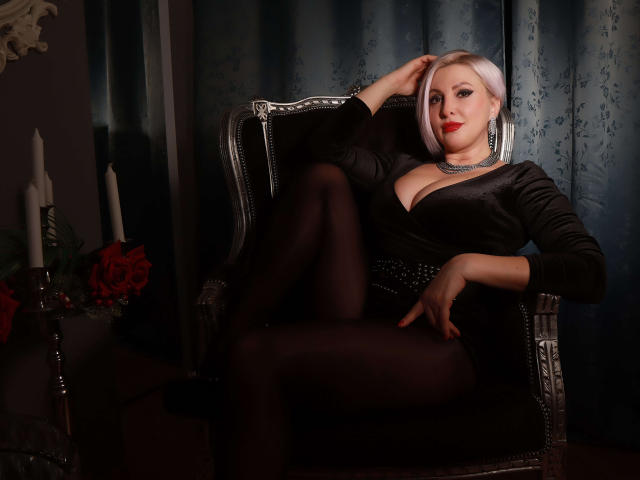 NicoleDomme with blonde bob, black dress, and cleavage, lounging on a silver chair, legs crossed, hand on head, black tights, red lipstick, and a necklace, looking sexy.