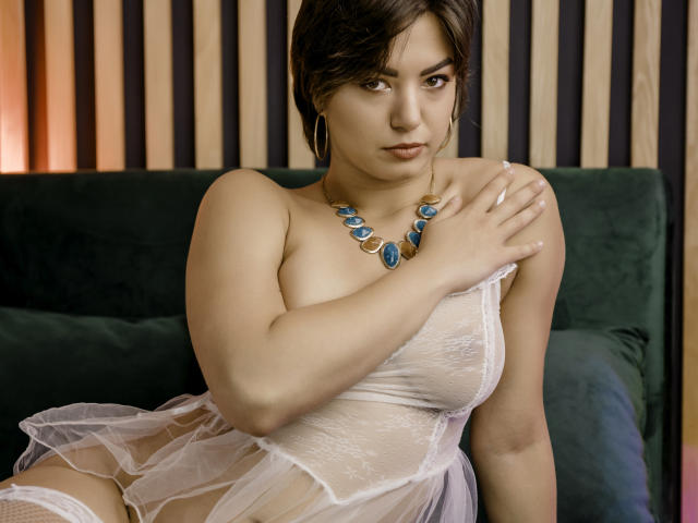 Monica Luiso, con un seno prosperoso, indossa un abito bianco trasparente, una collana con un gioiello blu, ha i capelli scuri, è seduta su un divano verde, ha una mano sul petto, guarda la fotocamera, illuminazione soffusa, sexy, seducente..