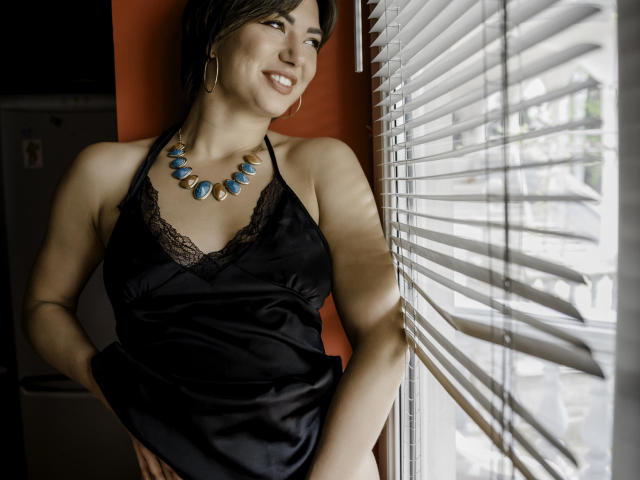 MonicaLuiso with brown hair, black lace halter top, and blue necklace, standing by window, smiling, showing her tits, wearing hoop earrings, sunlight through blinds, looking sexy.