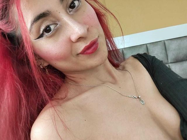 Victoria Millar hat rotes Haar, dunklen Eyeliner, roten Lippenstift, Sommersprossen, eine silberne Halskette und ein schwarzes Oberteil, das ihre nackten Schultern und einen Hauch ihrer Brüste zeigt..