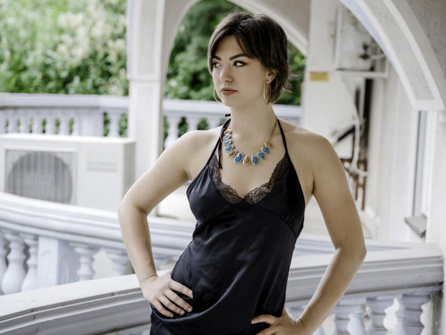 Monica Luiso, vêtue d'une robe en satin noir avec de la dentelle, un collier avec des pierres bleues, des cheveux bruns courts, les mains sur les hanches, regardant sur le côté, debout sur un balcon blanc, en journée..