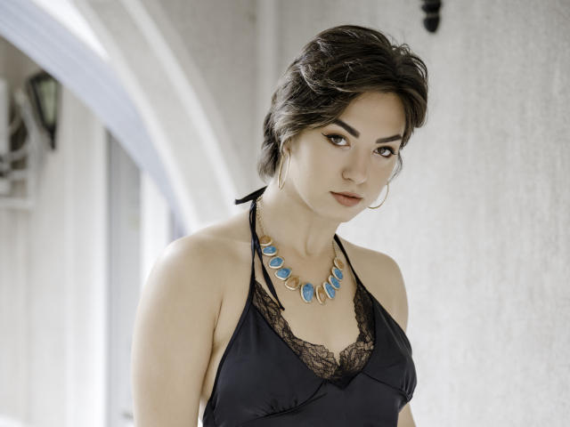 MonicaLuiso with short brown hair, wearing a black lace halter top, showing her tits, and a blue gem necklace, standing with a sultry look, indoors.