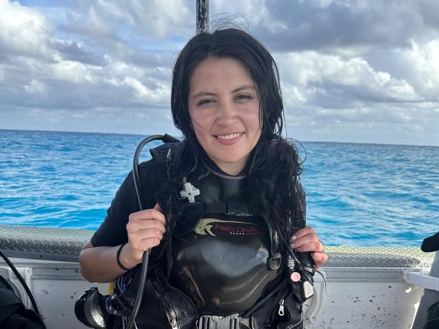 AmelieMuller with wet black hair, smiling, wearing a black scuba diving vest, holding a coiled rope, on a boat, blue ocean in background, cloudy sky overhead.