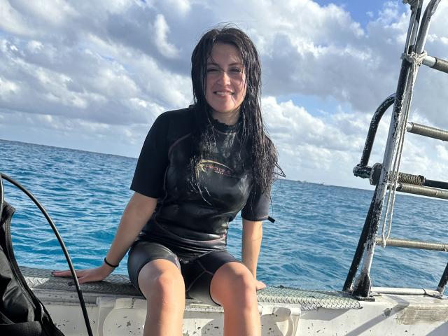 AmelieMuller, with wet black shirt and shorts, sits on a boat's metal deck, smiling, her long dark hair slicked back, blue ocean and cloudy sky in the background.