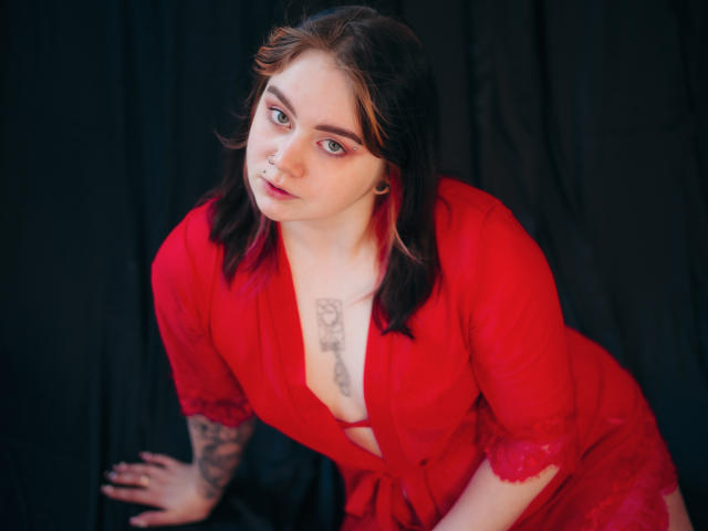 LunaLise in a red, sheer, button-down robe, leaning forward with a tattoo on her chest, has red lipstick, dark hair, and a tattoo on her right arm.
