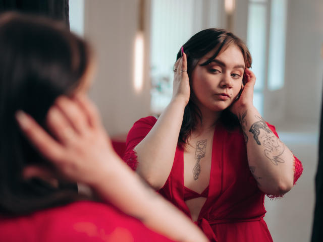 LunaLise with tattoos, red dress, and pink hair clip, adjusting her hair in front of a mirror, showing her tits and a small nose piercing, has a sexy look.
