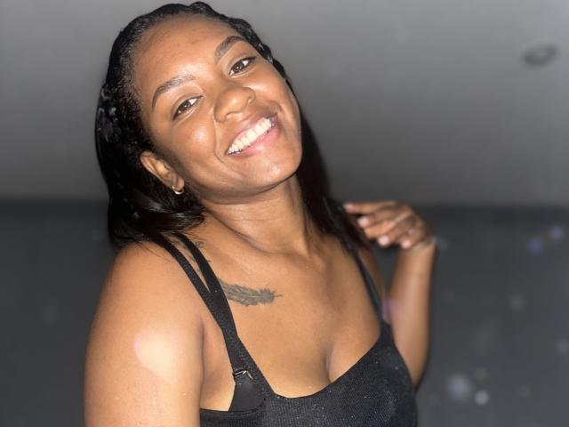 TiffanySammy, com pele escura, sorrindo, vestindo uma regata preta, mostrando seios pequenos, cabelo trançado preto, tatuagem no ombro direito, mão perto da orelha, com um brilho na foto..