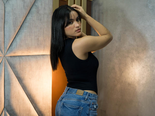 MiiaPalmer with black hair, wearing a black sleeveless top and blue jeans, standing with one arm raised, looking over her shoulder, showing her ass.