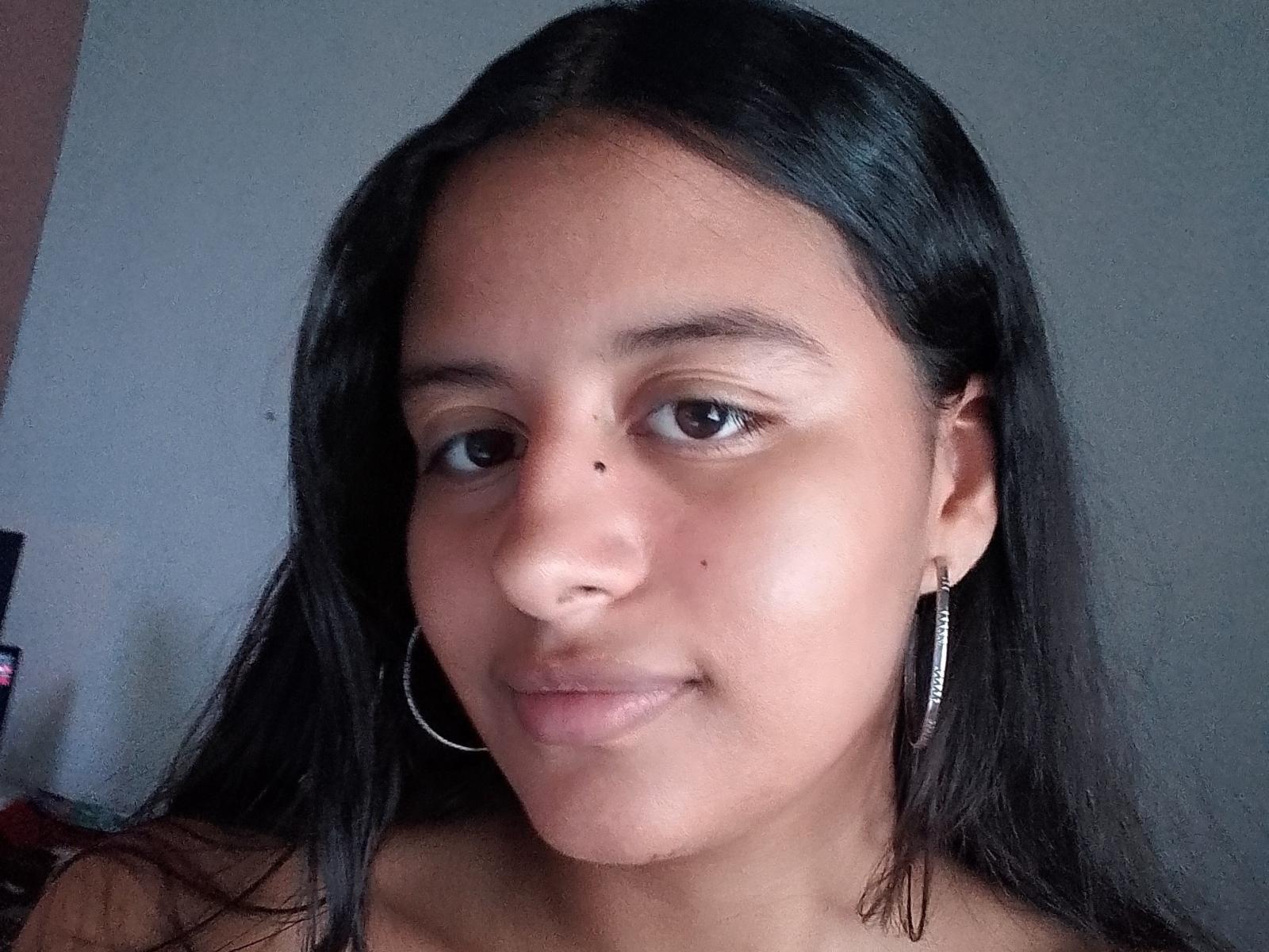BabeDollyS has long black hair, brown eyes, and a small mole on her right cheek. She's wearing thin hoop earrings and smiling slightly with no clothes visible.