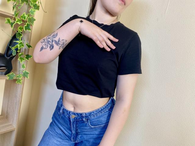 Swoiia with a black crop top, showing her flat stomach, blue jeans, and a floral tattoo on her right arm, her hand touching her chest.