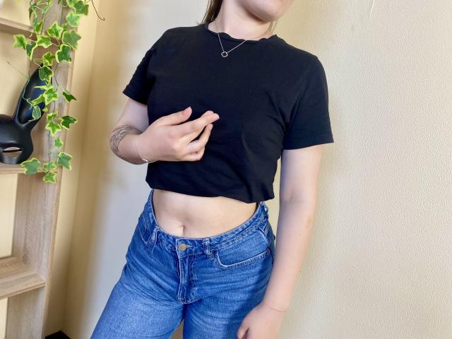 Swoiia with a black crop top, blue jeans, and a tattoo on her left arm, touching her flat stomach, wearing a necklace, with a green plant on the left.