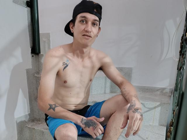 Tätowierter Mann ohne Hemd, mit schwarzer Kappe, in blauen Shorts, sitzt auf einer Betontreppe, schaut in die Kamera, zeigt ein leichtes Lächeln, die rechte Hand ruht auf dem Knie..