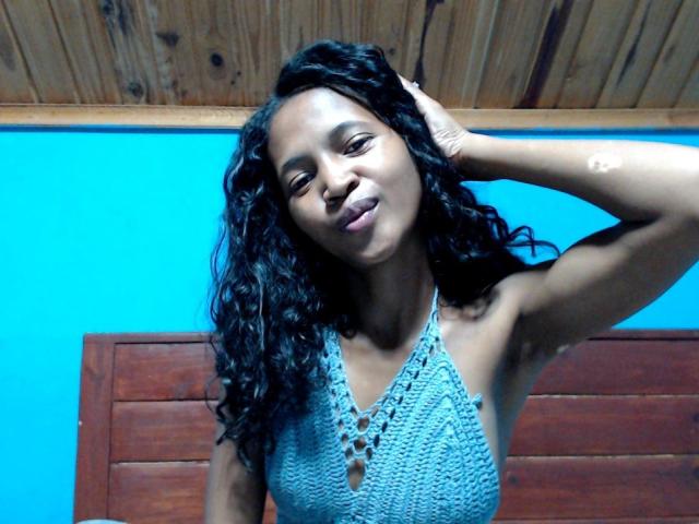 AmandaSexySexy with dark curly hair, wearing a blue crochet halter top, smiling slightly, hand in hair, wooden ceiling, and blue wall in background. Boobs visible through top.