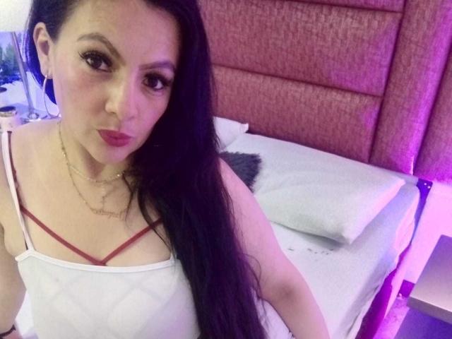 JakyKidman with long black hair, wearing a white camisole showing red bra straps, sitting on a bed with pink headboard, looking at the camera, purple light in background.