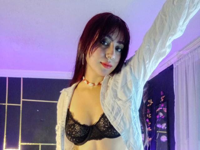 SarahLark mit roten Haaren, trägt einen schwarzen Spitzen-BH, ein offenes weißes Hemd und eine Halskette, Arm erhoben, blaues Augen-Make-up, Nasenring, lächelnd, violette und weiße Raumbeleuchtung..