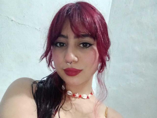 SarahLark has red and black dyed hair, a nose ring, and wears a red and white shell necklace. Her lips are painted red, and she has dark eye makeup.