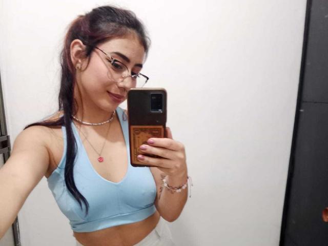 SarahLark mit Brille, hellblaues Sport-BH, zeigt kleine Brüste, rosarer Anhänger, langes, dunkles Haar, macht ein Selfie im Spiegel, hält ein Handy, weiße Wand, lächelt..