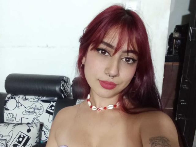 SarahLark, con i capelli rossi, un anello al naso e una collana di conchiglie, senza top, che mostra il seno, un tatuaggio sul braccio destro, rossetto rosa e eyeliner nero, seduta su una sedia nera..