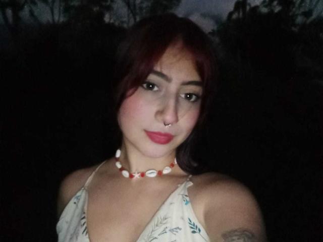 SarahLark tiene el cabello castaño con tintes rojizos, un piercing en la nariz, lápiz labial rojo y un collar de conchas marinas blancas. Lleva una blusa estampada y se puede ver un tatuaje en su brazo derecho..