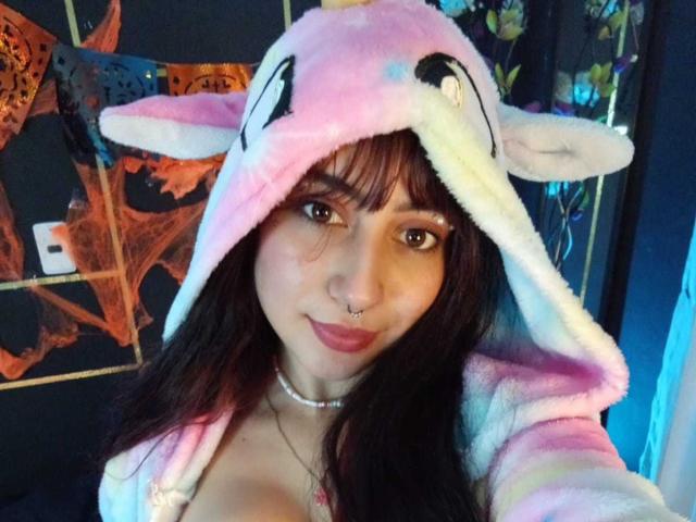 SarahLark with dark brown hair, pink nose ring, and a cute pink and white furry unicorn hood, showing off her small tits and smiling. She's wearing a choker.