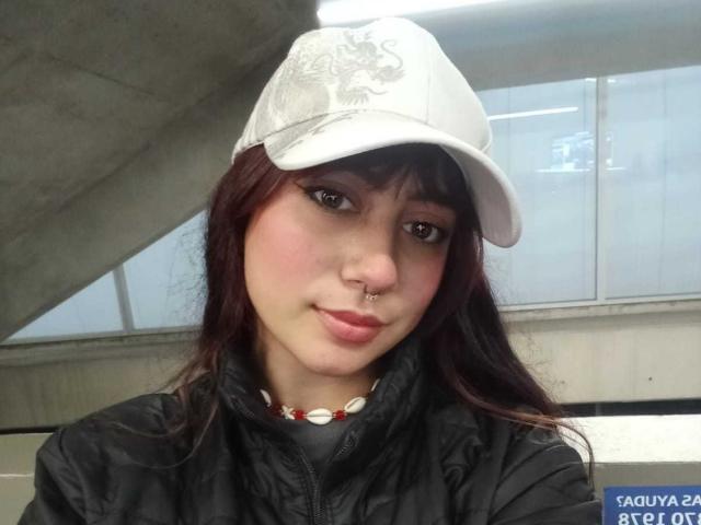 SarahLark, con cabello rojo oscuro, gorro blanco, chaqueta negra, piercing en la nariz y collar rojo, sonriendo ligeramente, mostrando su linda cara y sus labios rosados suaves..
