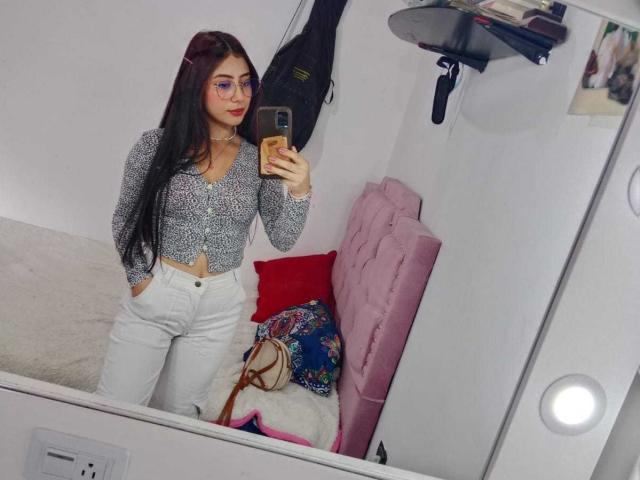SarahLark mit langem, schwarzem Haar, Brille und rotem Lippenstift, trägt ein gemustertes Oberteil und weiße Hosen und macht ein Selfie im Spiegel, wobei ihre kleinen Brüste und ihre Hand in der Hosentasche zu sehen sind..