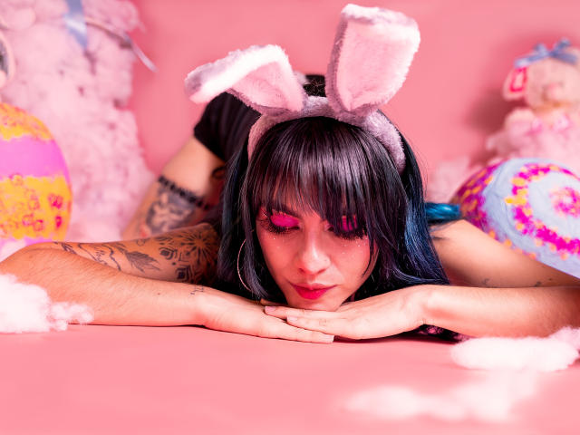 SamHazell with pink bunny ears, black hair, and tattoos, face down on pink floor, hands under chin, pink eyeliner, blue highlights, pink fluffy clouds, colorful stuffed toys.