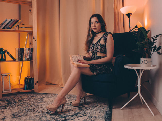 Decadance with long brown hair, wearing black lace lingerie, sits in a black chair, reading a book, in nude heels, with a choker, on a patterned rug.