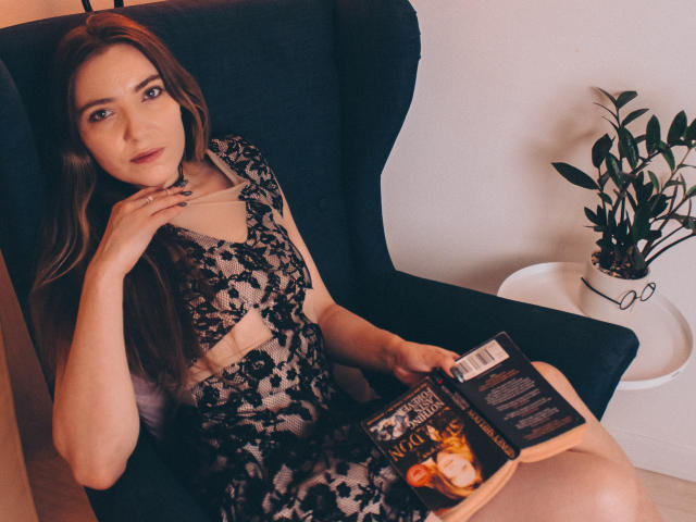 Decadance with long brown hair, lace dress, and blue eyes, holding a book, sitting in a dark chair, right hand on chest, left hand on book, potted plant beside.