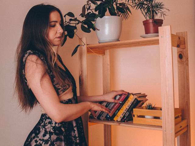 Decadance with long brown hair, wearing a black lace dress, holding books on a wooden shelf, looking down, next to potted plants. Her tits are small, her pussy is unseen.
