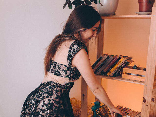 Decadance with long brown hair, black lace bra, and matching skirt, bending over a wooden shelf, touching books, has a cute butt and small, perky tits.