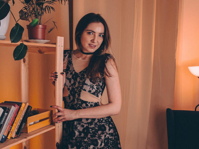 Decadance stands near a wooden shelf, wearing a black lace dress with cutouts, exposing her tits. She has long brown hair, a black choker, and a slight smile.
