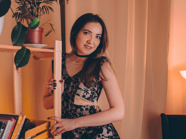 Decadance with brown hair, wearing black lace dress, holding wooden shelf, smiling, black choker, beige curtains, warm light, potted plant, books, and yellow shelf in background.