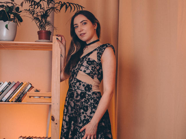 Decadance stands in a room wearing a black lace dress with cut-out sides, a black choker, and has a cute smile. She's next to a wooden shelf with books.
