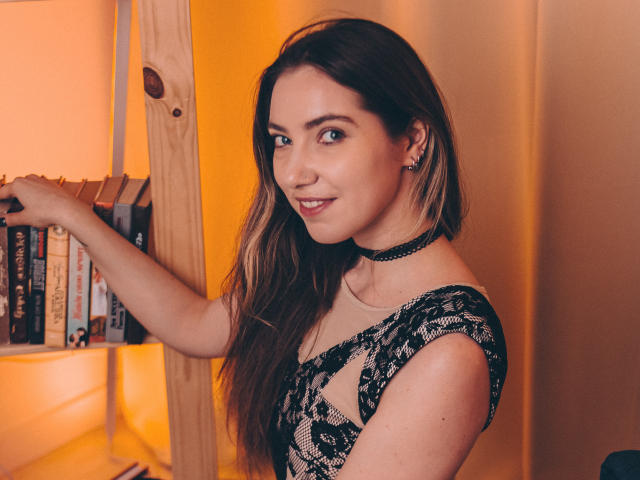 Decadance with long brown hair, lace top, and black choker, standing by a wooden bookshelf, smiling, showing her tits through lace, holding books. Warm yellow light.