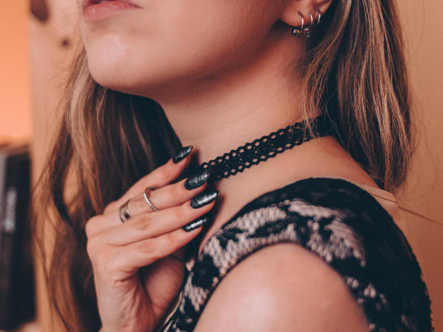 Decadance's face, showing her neck and hand, with black lace top, black choker, and glossy black nails, wearing a silver ring and hoop earring, in a soft light.