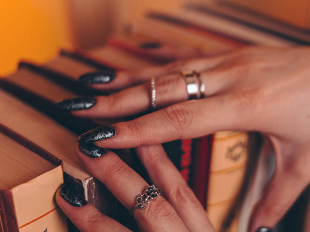 Decadance's hands with black, glittery nails and multiple rings are on a stack of books. Her fingers are slightly curled, showing off her shiny, decorative nail art.