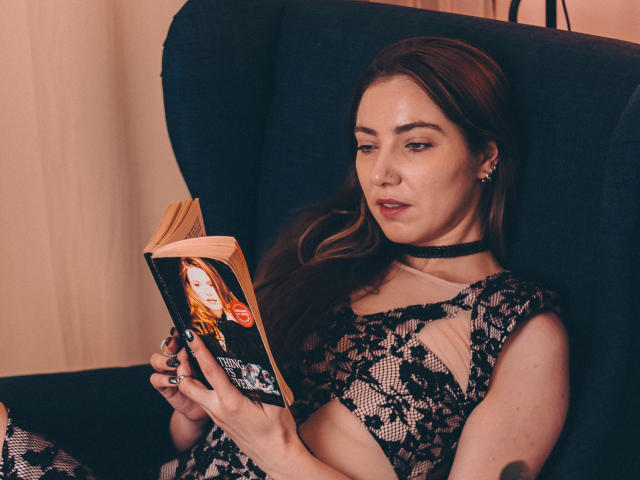 Decadance with long brown hair, wearing black lace dress and choker, reading book with red-haired woman on cover, sitting in dark blue chair, looking focused.
