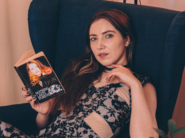 Decadance with long brown hair, blue eyes, wearing black lace dress, reading "Spell Dusk: Witch of Endor" book, sitting in blue armchair, hand on chest, slightly seductive look.