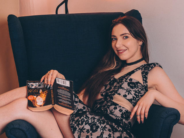 Decadance with long brown hair, smiling, wearing black lace lingerie, sitting in a blue chair, holding a book with a flame design, and a black choker.