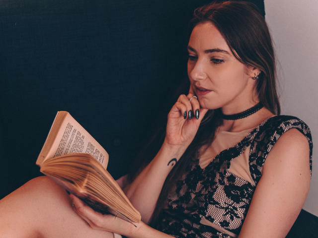 Una donna con lunghi capelli castani, una maglia di pizzo nero e un collare, che legge un libro, tenendolo con una mano e toccandosi le labbra con l'altra, con unghie scure e un piccolo tatuaggio sul polso..