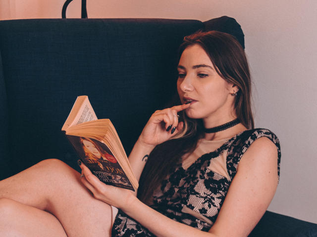 Decadance with long brown hair, lace dress, black choker, and black nail polish, reading a book, finger in mouth, legs crossed, sitting on dark chair.
